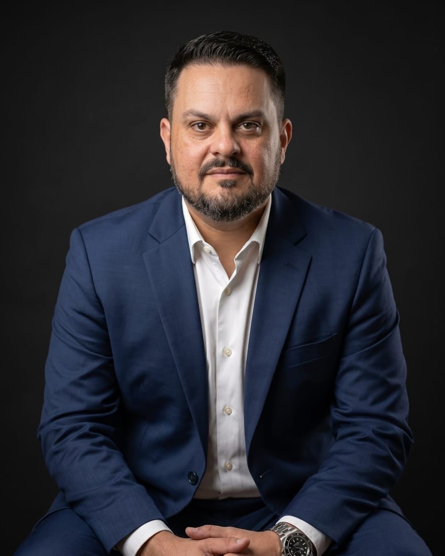 Cristhian Sanches de Souza, fundador da Virtus Mordomia, sentado, de terno azul, em fundo escuro